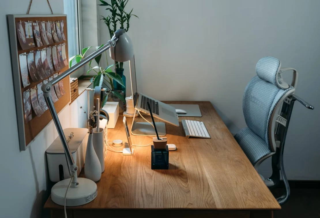  A desk with a laptop, keyboard, mouse, lamp, plants, and other office supplies on it, and an ergonomic chair behind it.